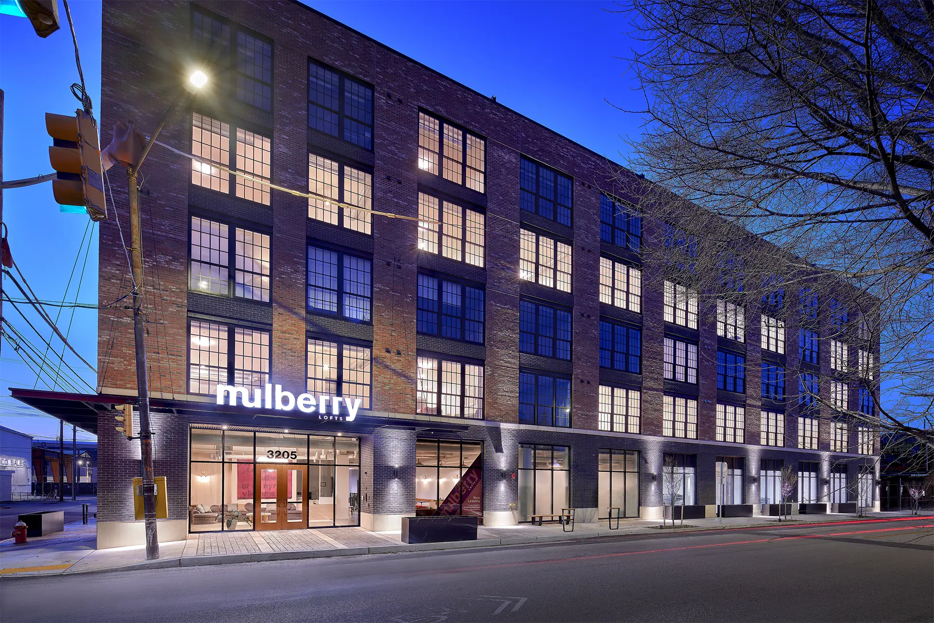 Exterior of a modern brick apartment building at dusk, featuring large industrial-style windows and a street-level lobby entrance